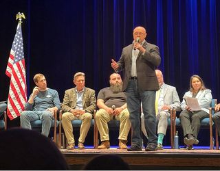 A man in a suit talking into a microphone on stage, while a row of other people sit behind him along with an American flag